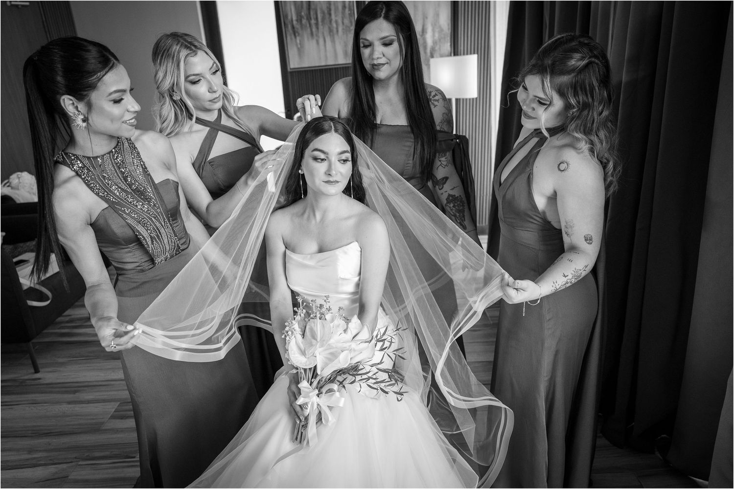 Wedding preparation scene with bridesmaids helping adjust the bride's veil in black and white.