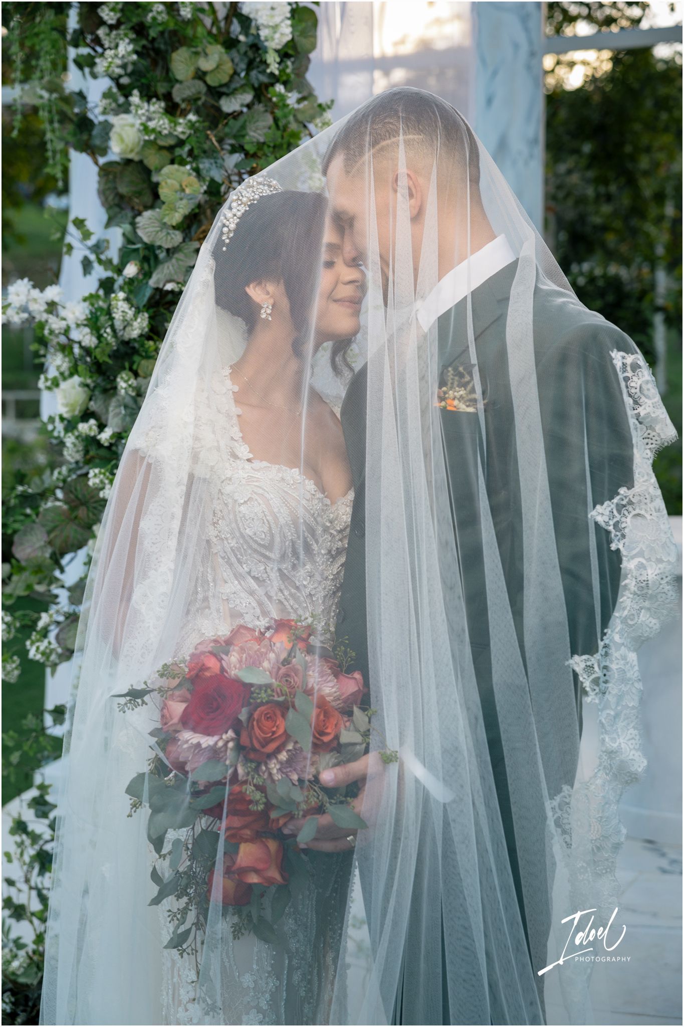 Wedding couple shares an intimate moment under flowing lace veil with red rose bouquet and floral arch backdrop.