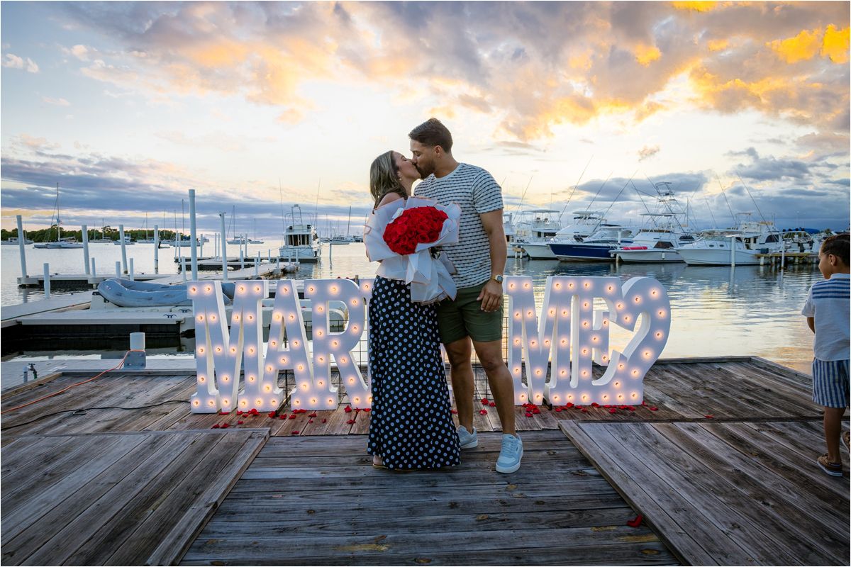 A romantic proposal setup on a marina dock at sunset with lit LOVE letters and boats in the harbor background.