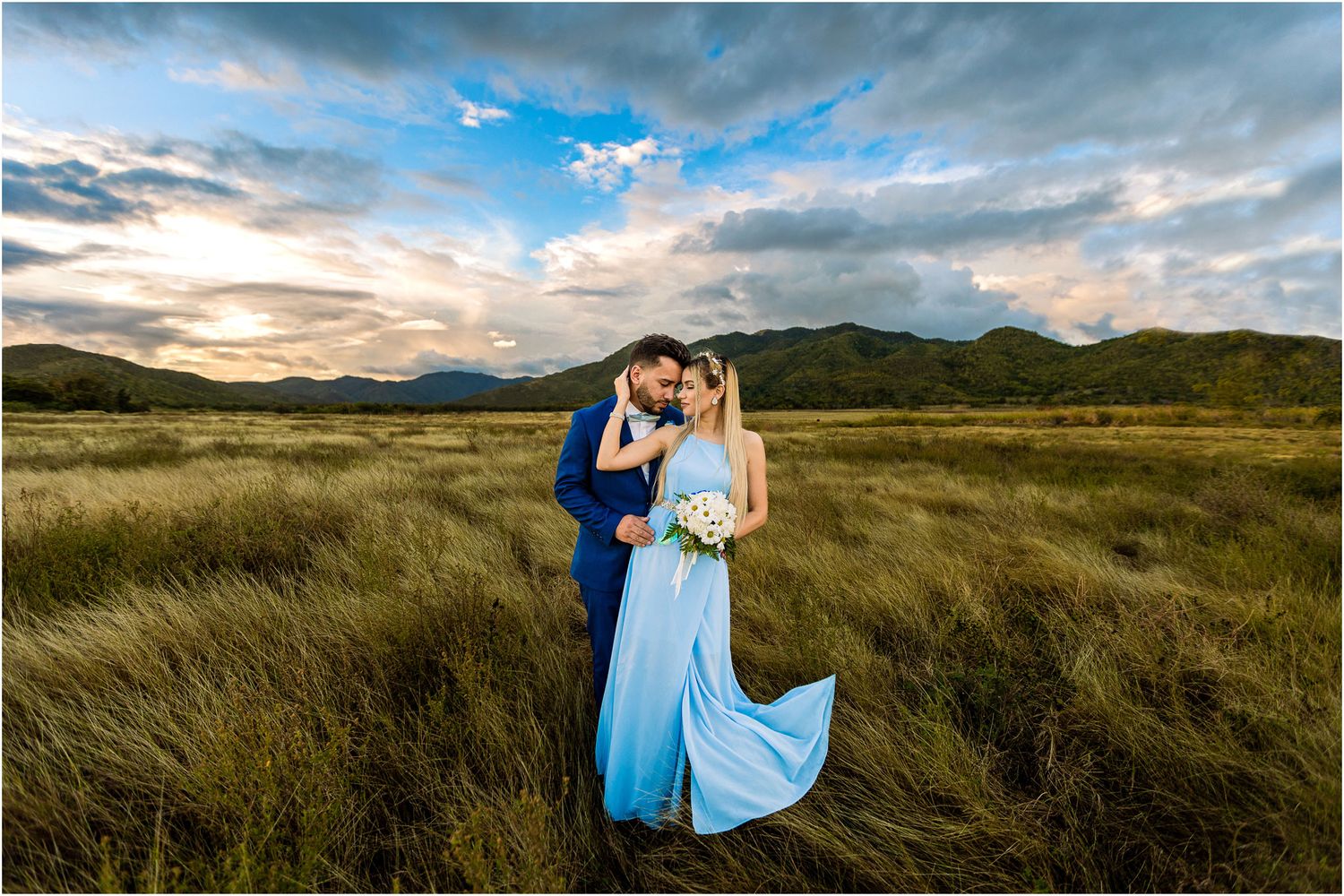A couple in blue formal wear stands in a windswept field with dramatic sunset sky and mountains.