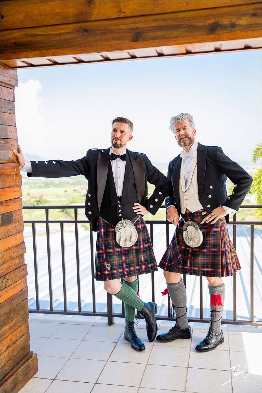 Two groomsmen in traditional Scottish kilts and formal attire pose on a balcony with scenic views.