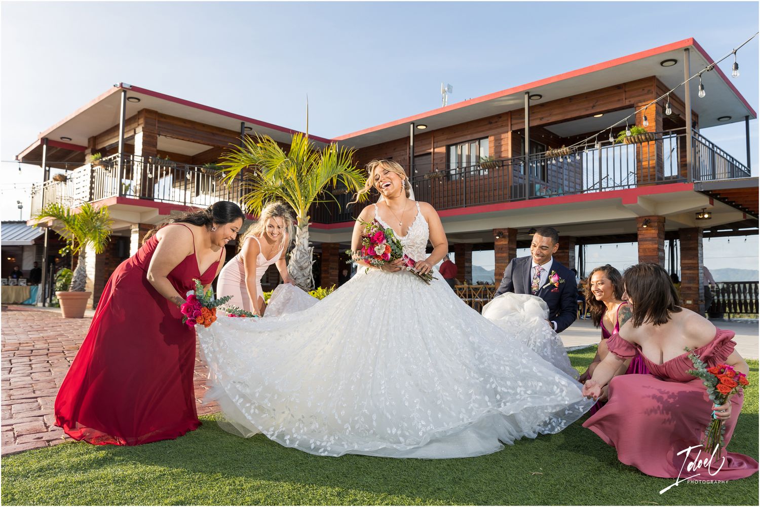 Bridal party in burgundy dresses arrange white wedding gown on lawn in front of modern venue.