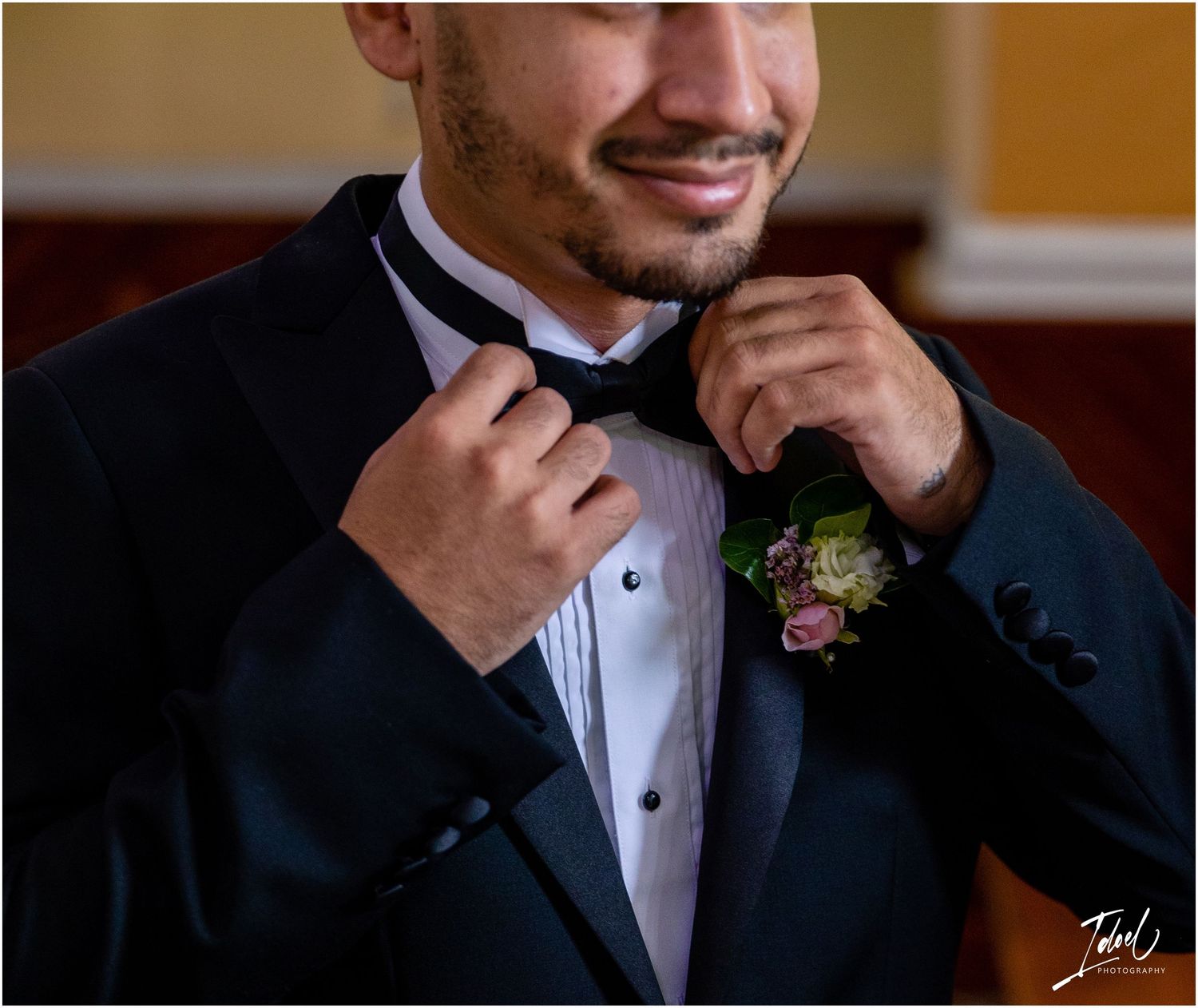 Candid moment of groom adjusting bow tie while wearing classic black tuxedo with floral boutonniere.