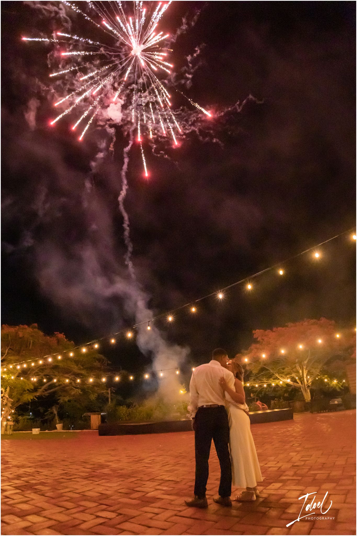 Romantic moment under bursting fireworks with string lights illuminating the outdoor venue.