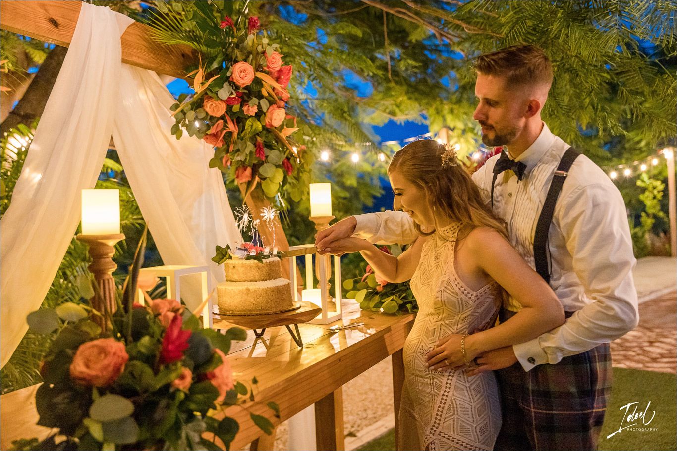 Couple shares intimate moment while cutting their wedding cake under string lights and floral arch.