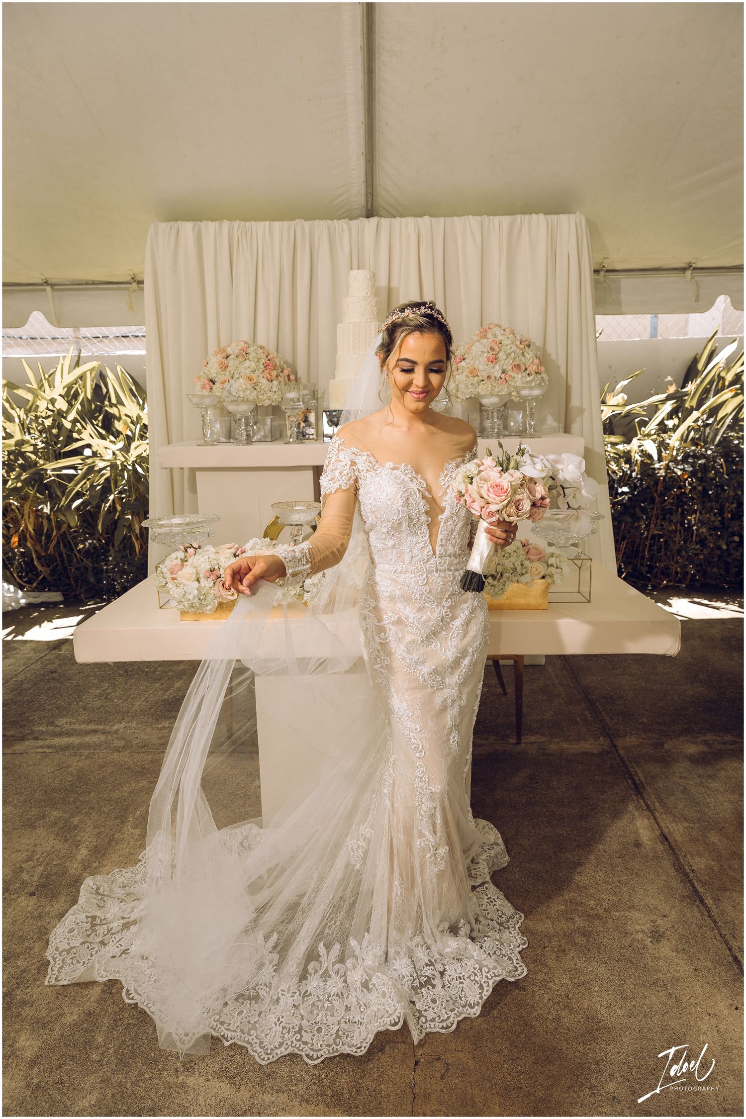 Elegant bride in lace mermaid wedding dress holding pink bouquet against cream backdrop.