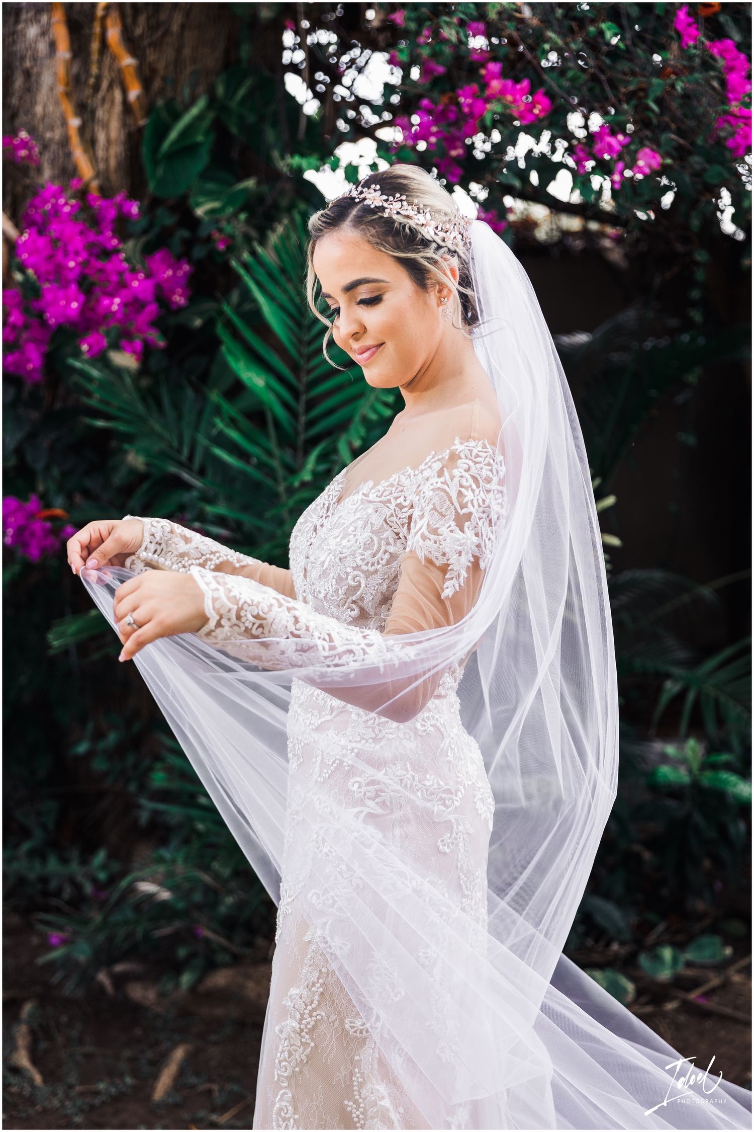 A bride in a lace wedding dress poses with her flowing veil against pink bougainvillea flowers.