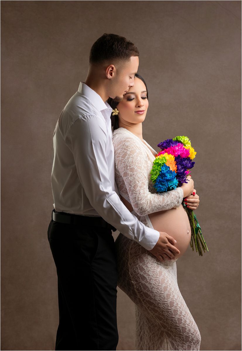 Romantic maternity photo series with couple in formal wear holding vibrant rainbow bouquet against neutral backdrop.