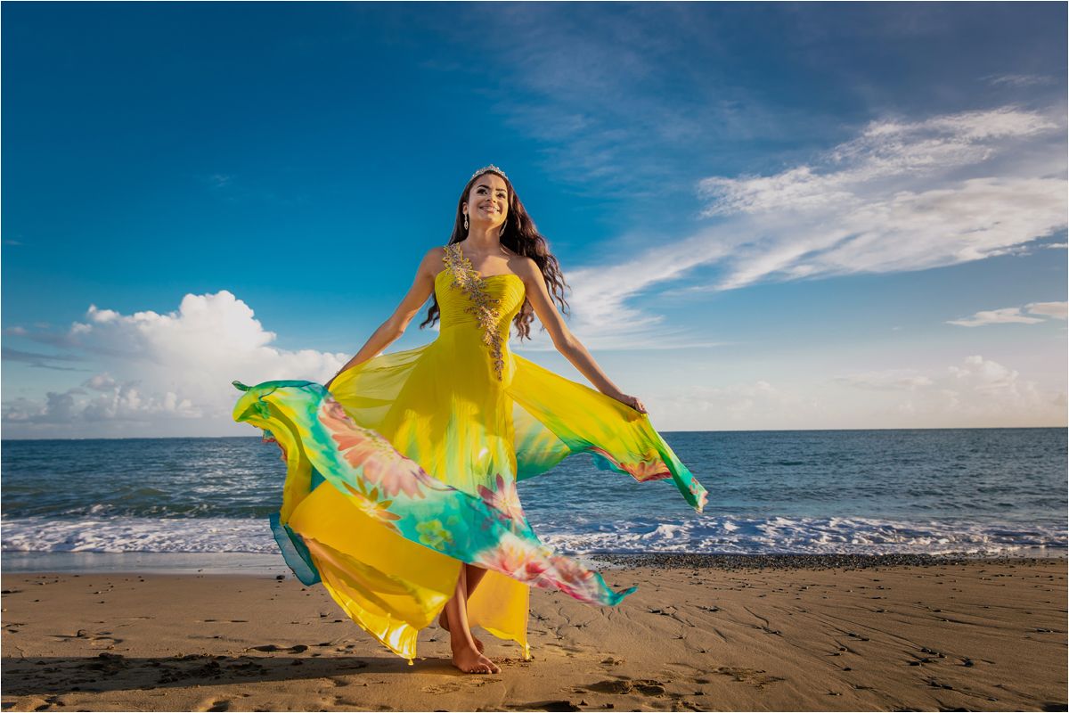 Person in flowing yellow dress twirls on beach against vibrant blue sky and ocean.
