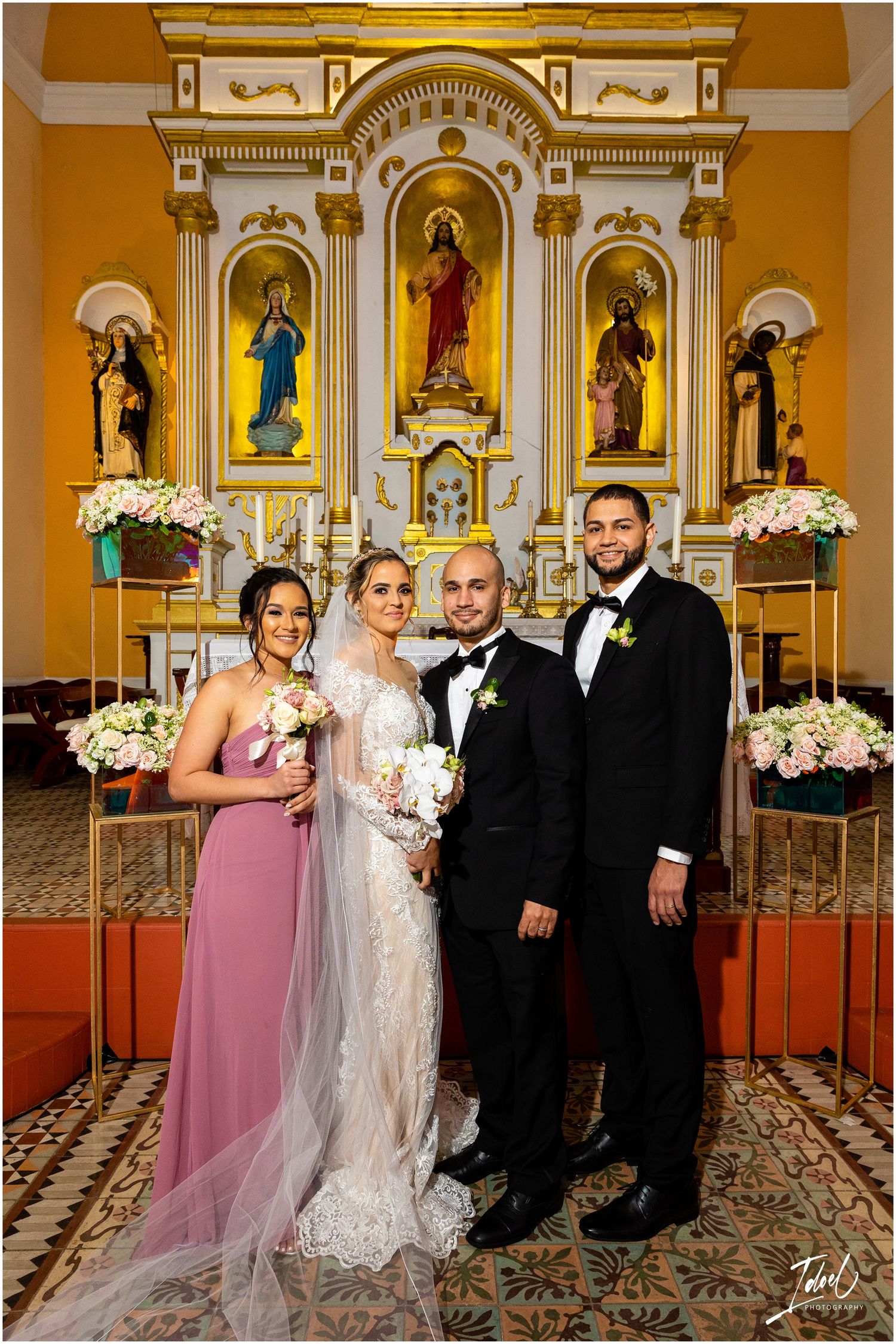 Wedding couple and attendants pose formally in front of ornate golden church altar with religious statues.