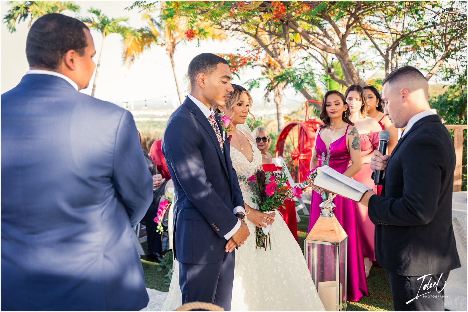 Outdoor wedding ceremony with bridesmaids in magenta dresses under flowering trees.
