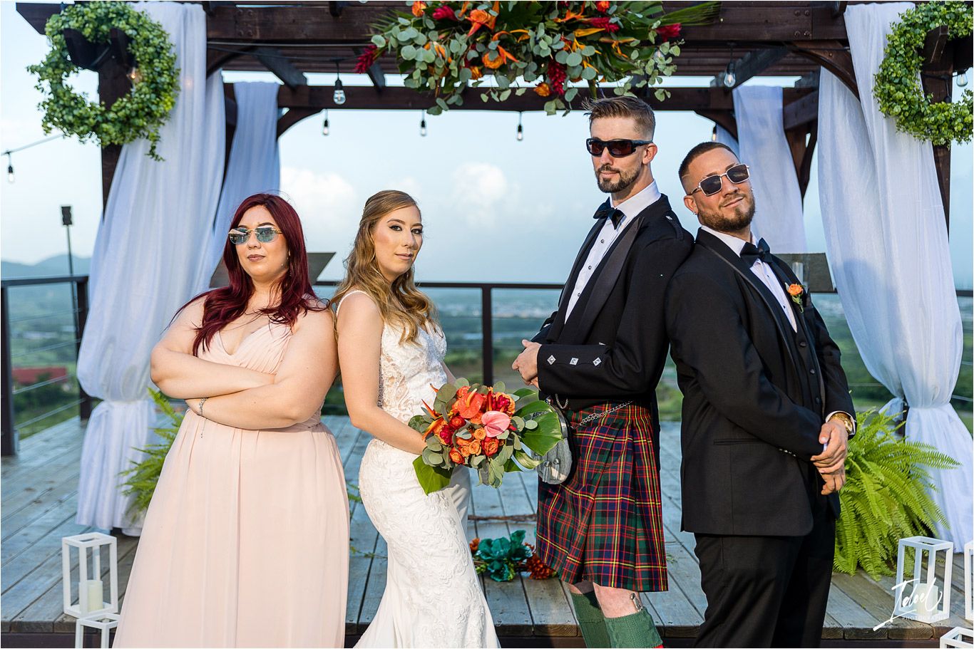 Wedding party poses outdoors under decorated pergola with white drapes and floral accents.