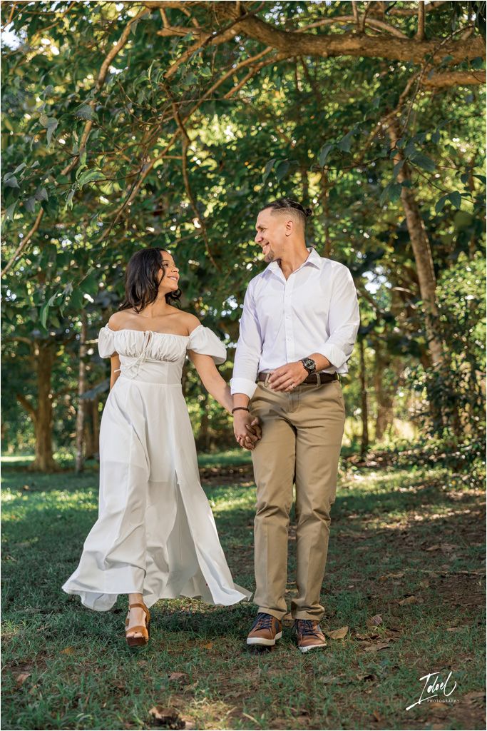 A romantic couple in casual attire holding hands and enjoying a stroll under tree branches in a garden setting.