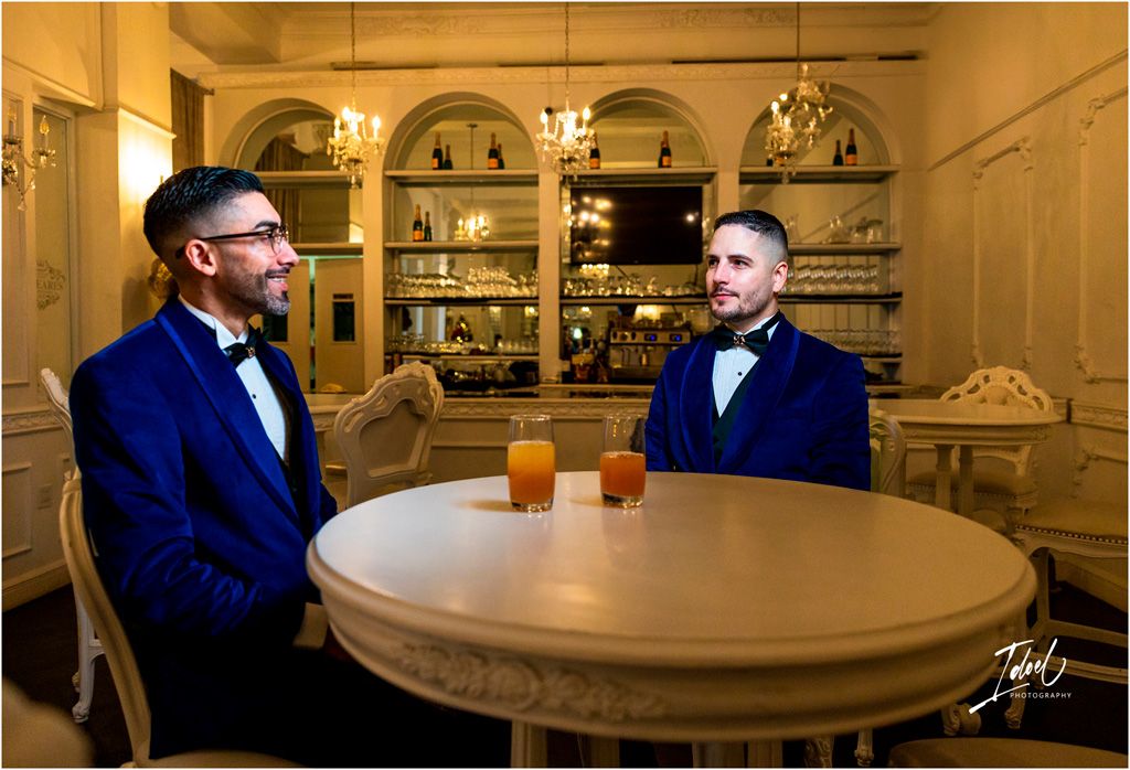 Two men in navy blue suits and bow ties sitting at a round table in an elegant restaurant interior with arched alcoves.