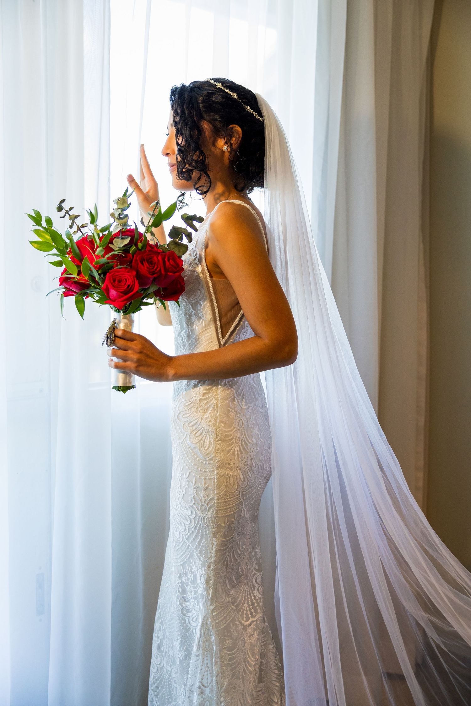 Bride in lace dress holding red bouquet by window with flowing veil.
