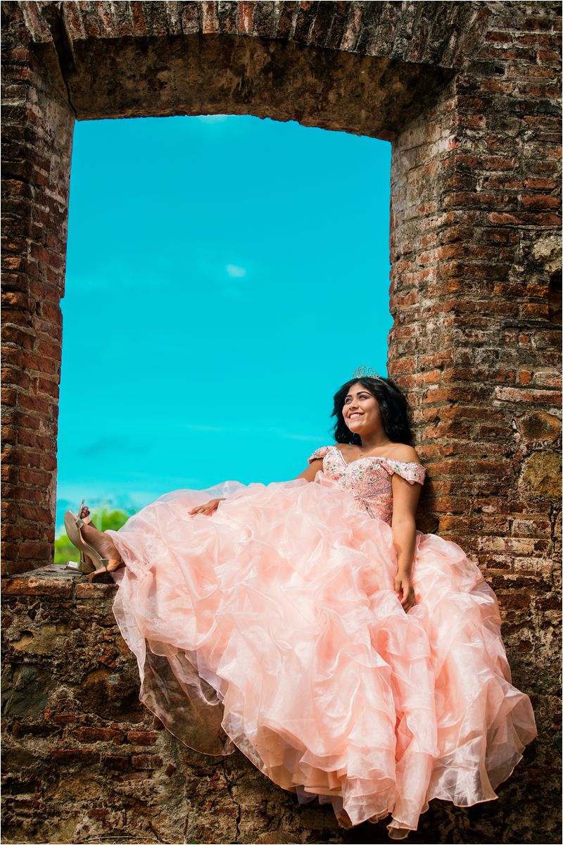 A person in a flowing peach-colored dress poses against a brick wall under bright turquoise sky in a photo series.