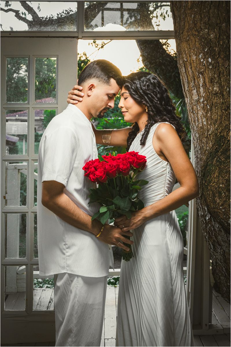 A romantic couple in white outfits shares tender moments while holding red roses against a rustic outdoor backdrop.