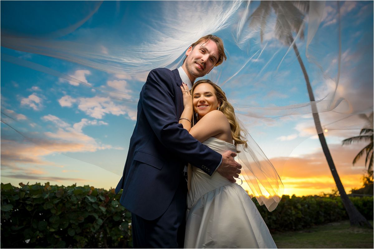 Newlywed couple embraces during sunset at a tropical beach wedding with palm trees and vibrant orange sky.