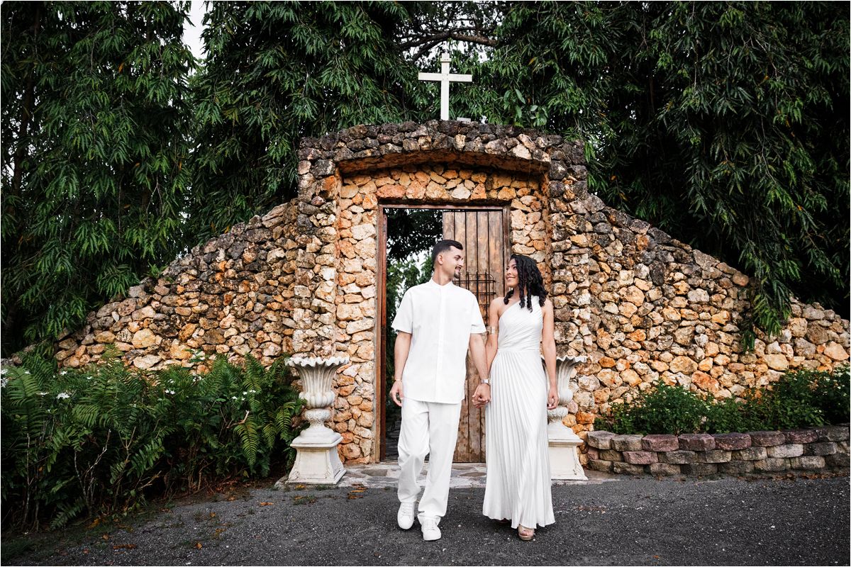 A couple in white outfits stands before a rustic stone grotto with a cross on top surrounded by greenery.