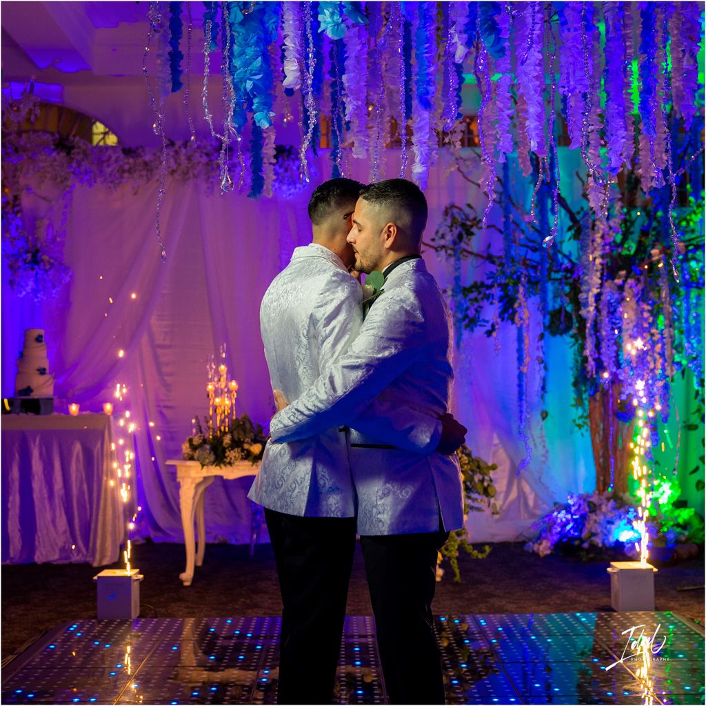 Two people embrace on a dance floor surrounded by dramatic blue and purple lighting and hanging decor at an elegant event space.