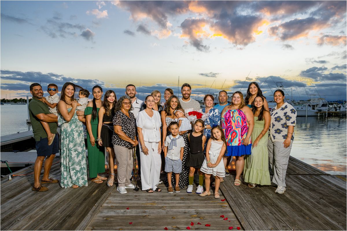 A large family group poses together on a wooden dock at sunset with a marina and colorful sky in the background.