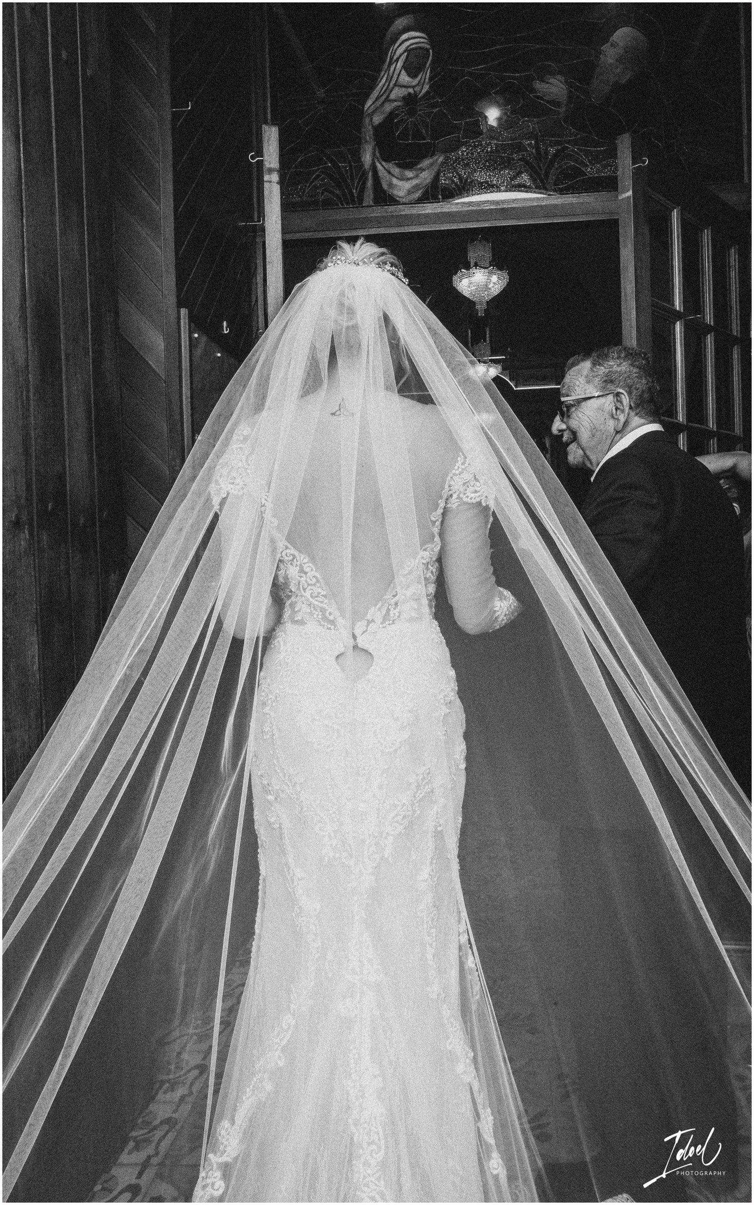 A black and white photo shows a bride in a lace dress with long veil walking down the aisle.