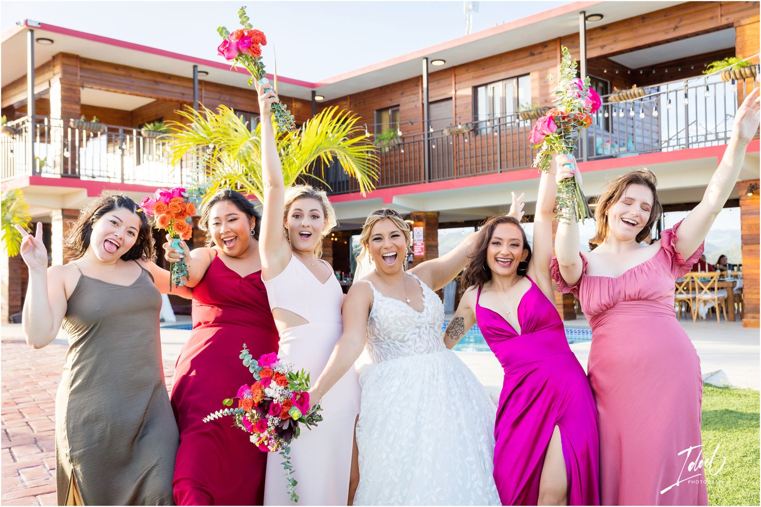 Wedding party in colorful dresses celebrates joyfully outside modern tropical venue.