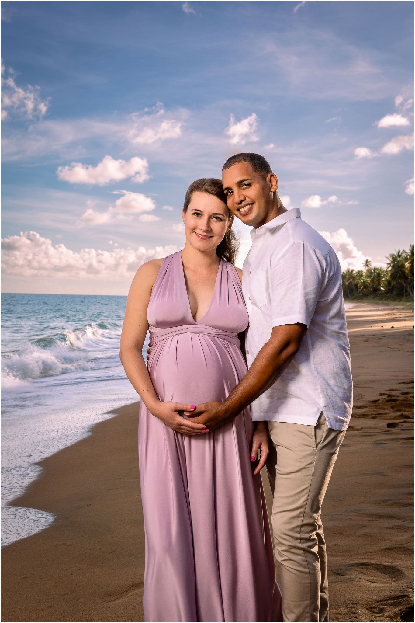 Maternity beach portrait of couple embracing on sandy shore with ocean waves and palm trees.