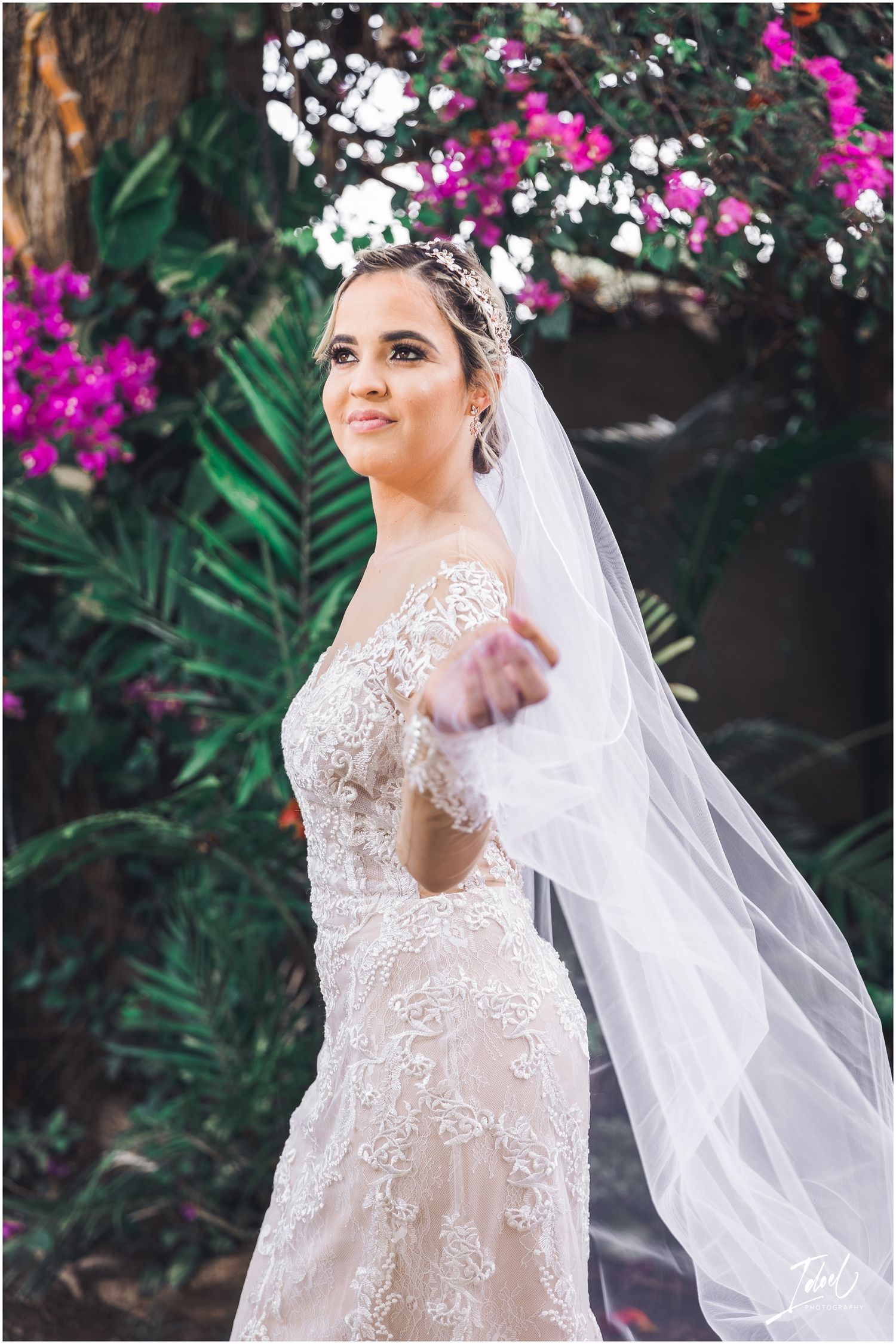 A joyful bride in a detailed lace dress poses with her veil against flowering bushes.