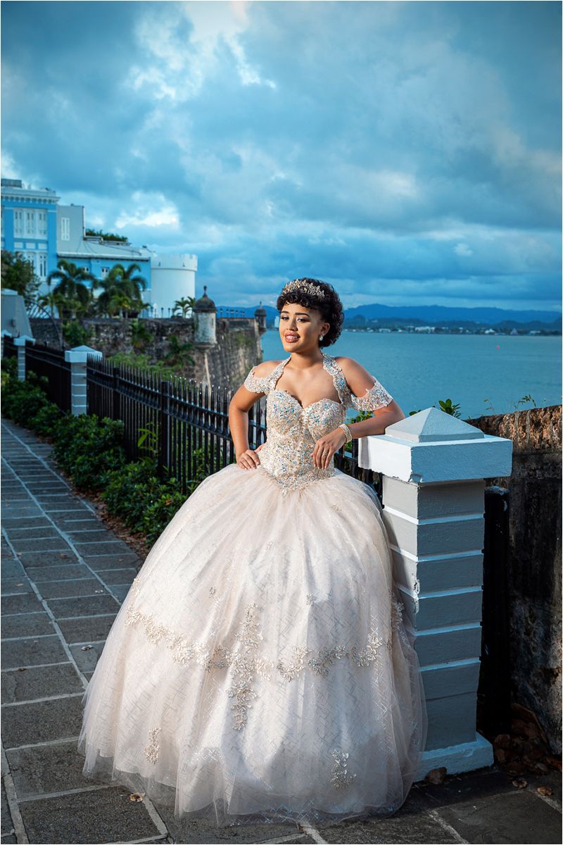 A person in an elegant white ballgown poses along a seaside wall with ocean and mountains in the background.