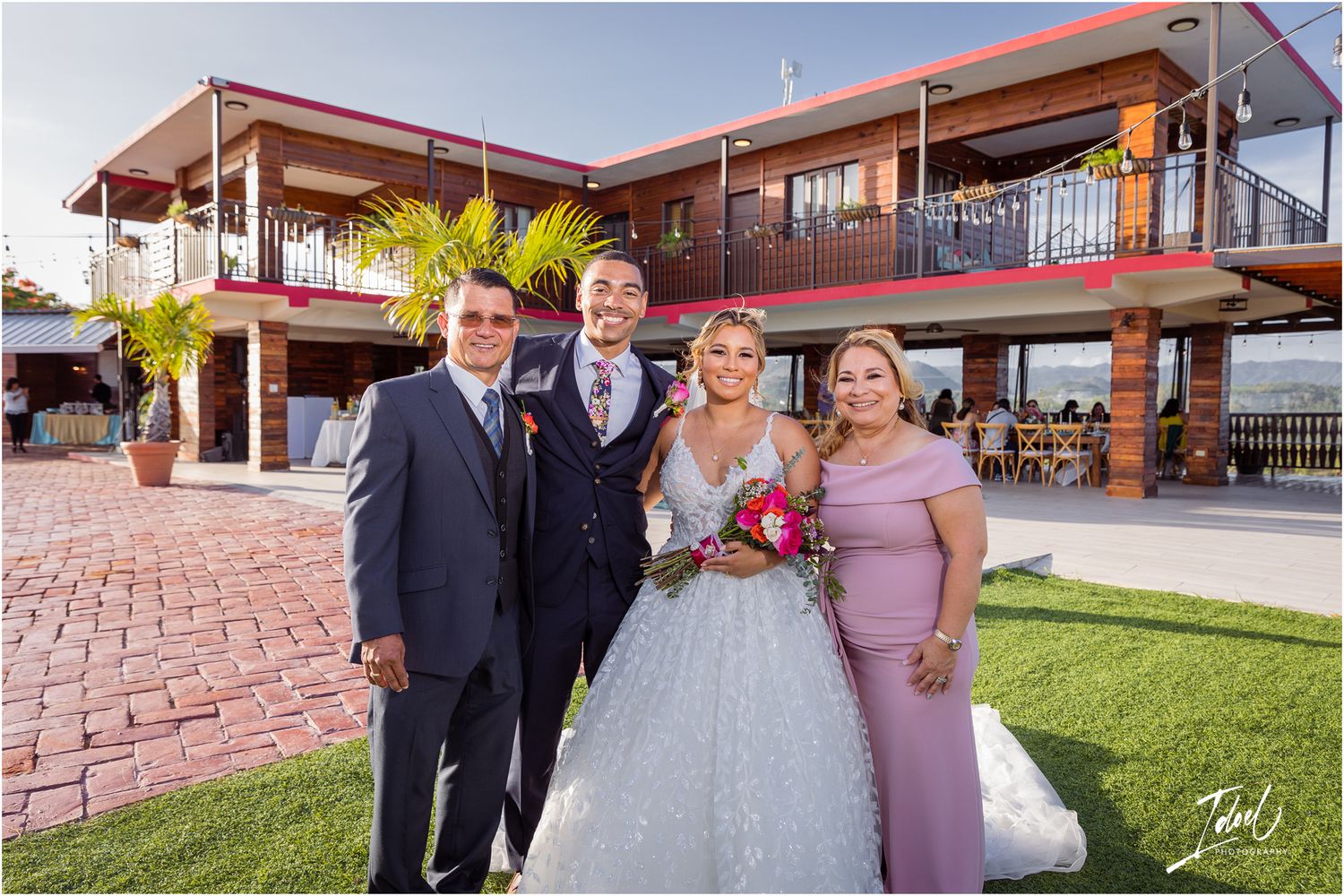 Wedding party poses in front of a modern two-story venue with brick pavers and palm trees.