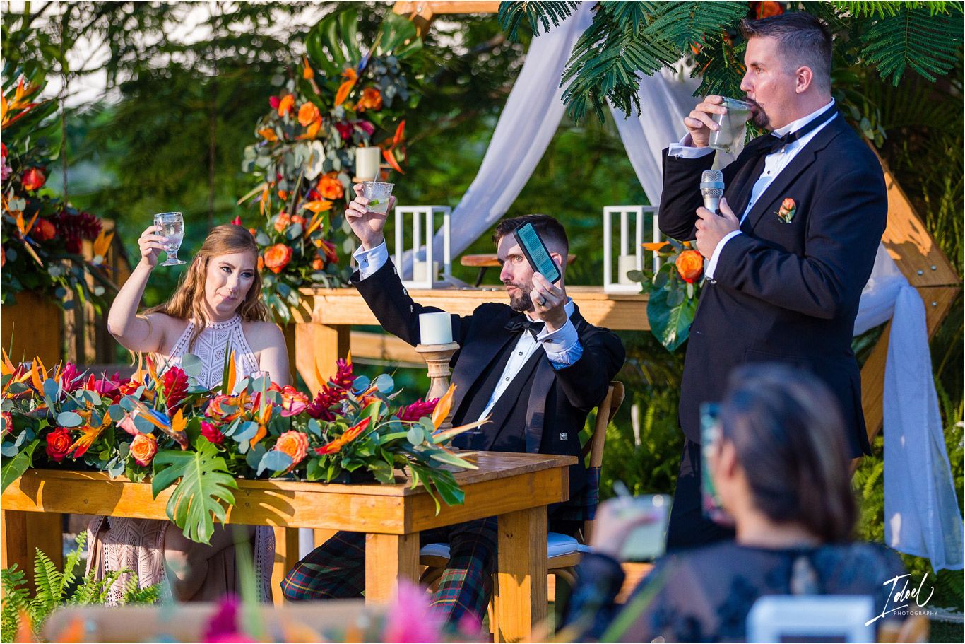 Wedding guests react joyfully during outdoor evening reception with floral decor and string lights.