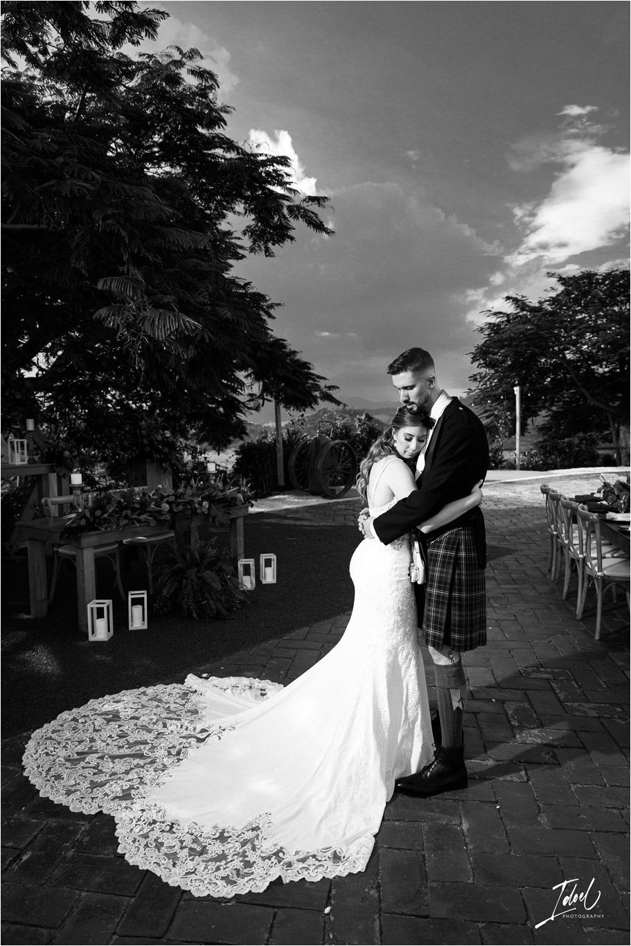 A black and white portrait of newlyweds with a dramatic lace train on an outdoor terrace.