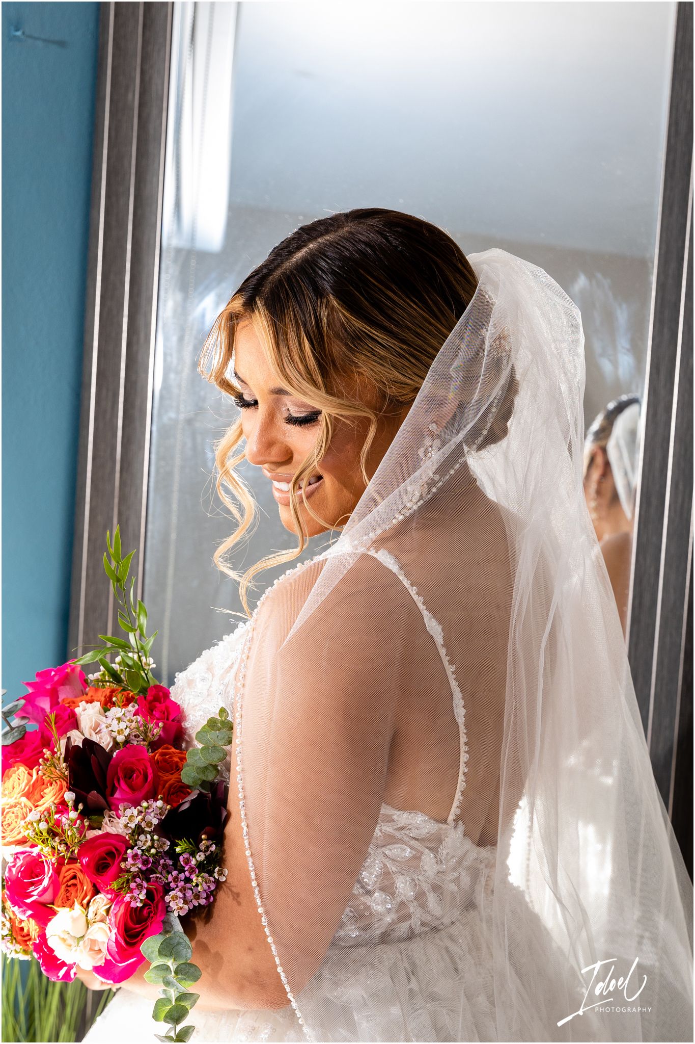 Elegant bridal portrait featuring a lace gown, veil, and vibrant pink bouquet by a window.