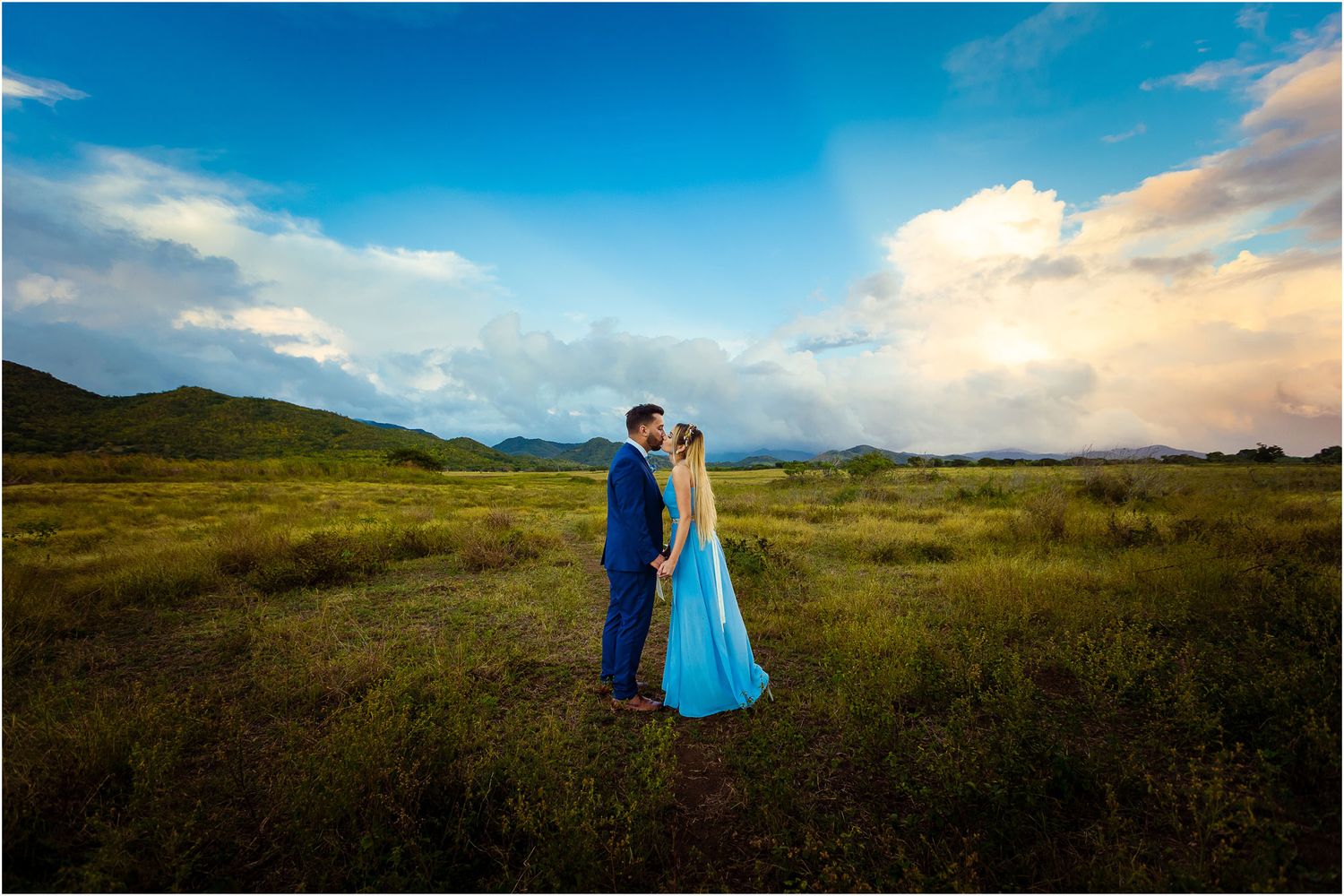 Two people stand close together in formal wear against a dramatic landscape of rolling hills and cloudy skies.