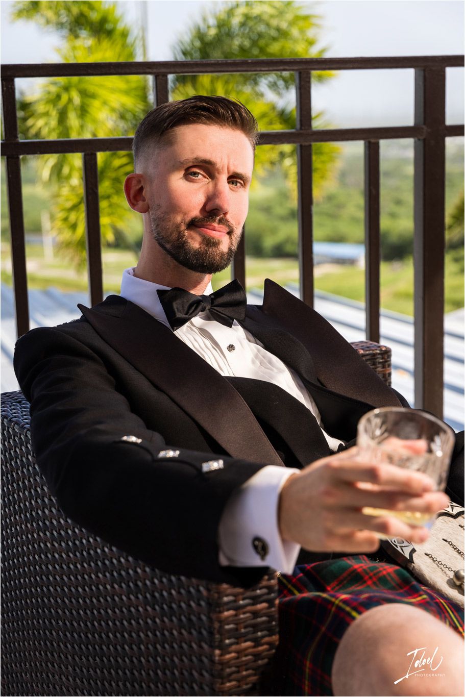 Groomsman in tuxedo and kilt relaxes with drink on outdoor patio furniture.