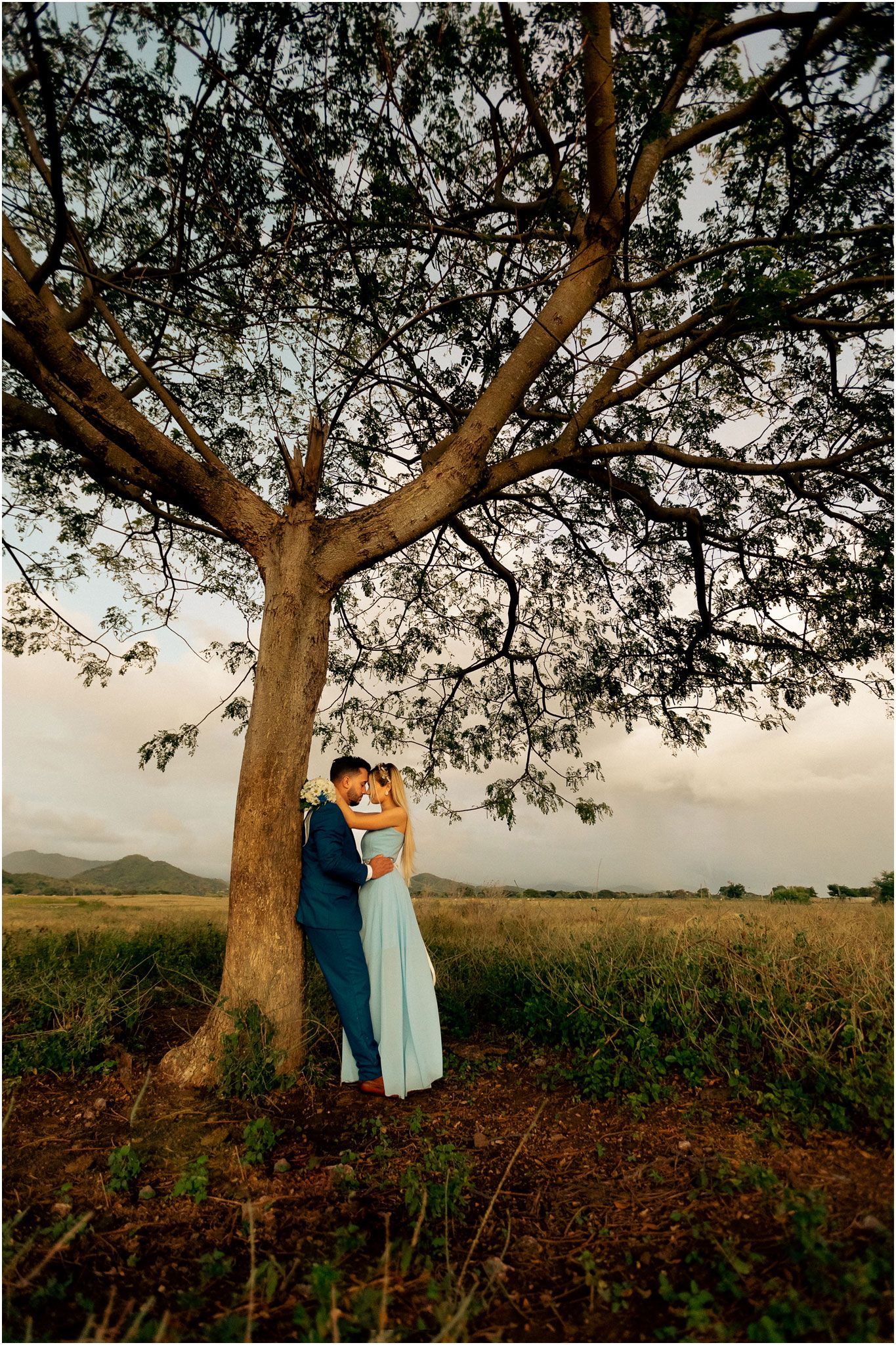 A couple embraces under a large tree during sunset in a rural field with mountains in the distance.