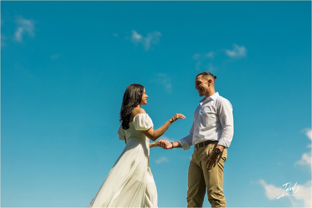 A couple in casual wedding attire stands together against a vibrant blue sky on a sunny day.