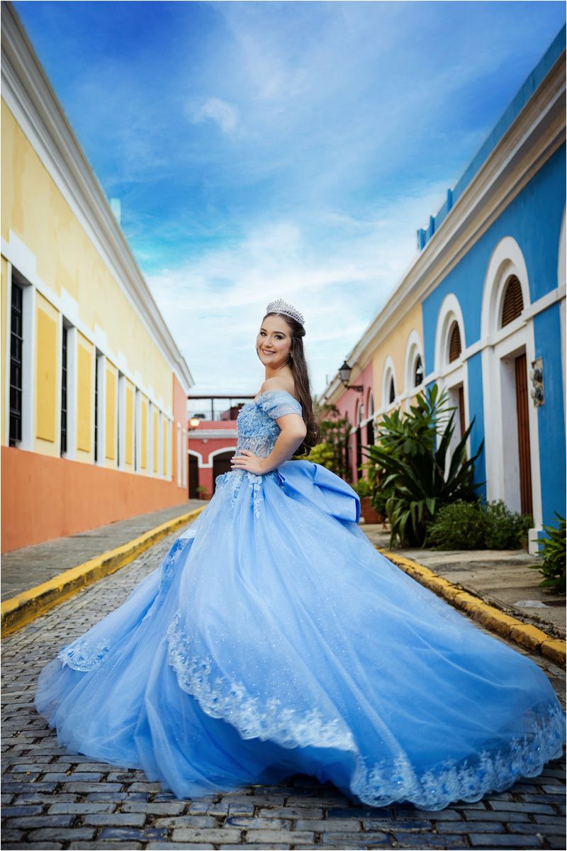 A stunning light blue ballgown dress flows across a historic cobblestone street with colorful colonial buildings in the background.