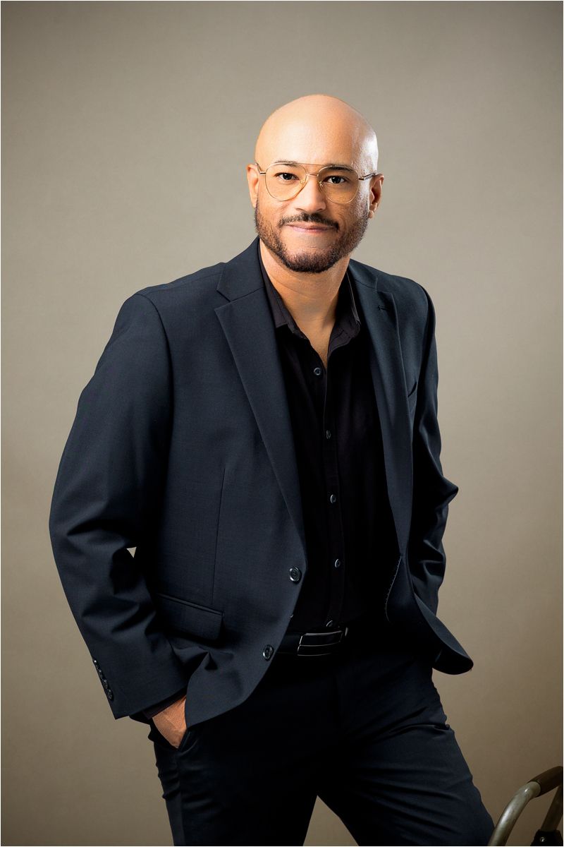 Professional portrait of someone in a dark suit and black shirt against a neutral background.