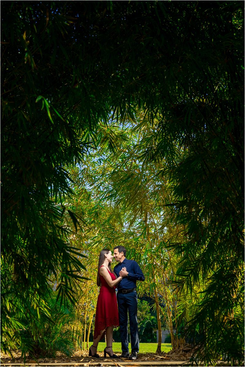 Couple sharing a romantic kiss framed by lush bamboo trees in a sunlit garden, woman in red dress.