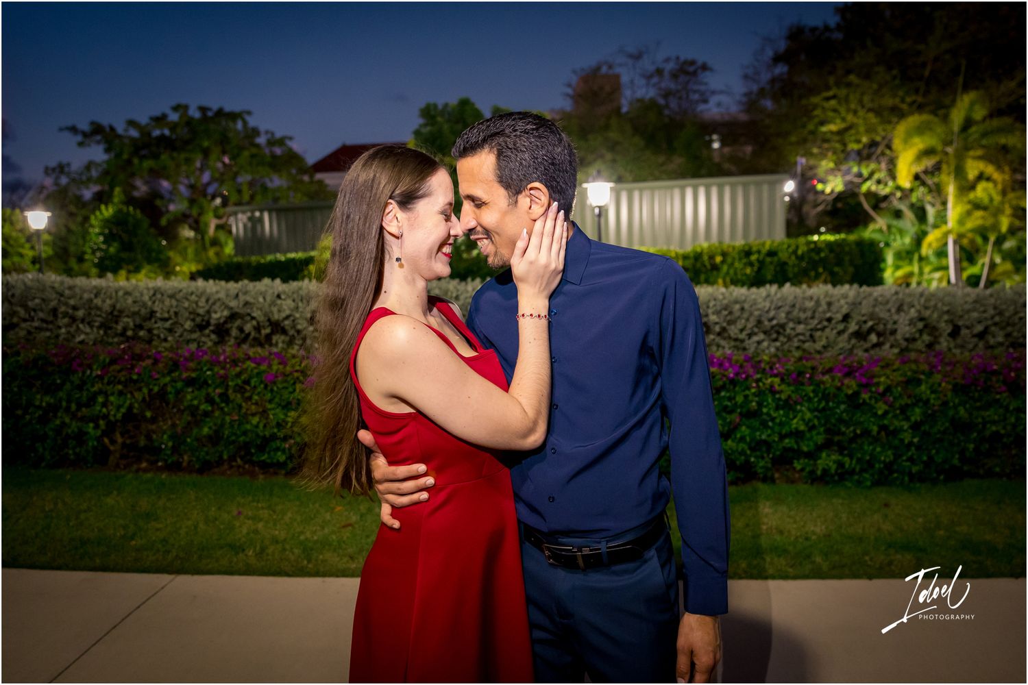 Romantic couple in red dress and blue shirt sharing an intimate moment in a lush garden at night.
