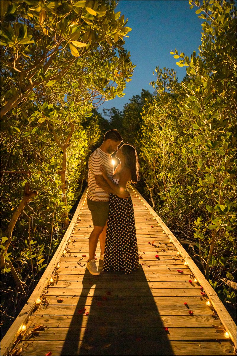 Silhouetted couple share romantic moment on illuminated wooden boardwalk through mangroves at night.