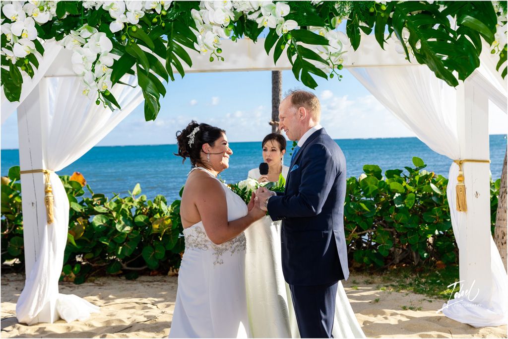 A beachside wedding ceremony under a white draped arch with tropical greenery and flowers overlooking the ocean.