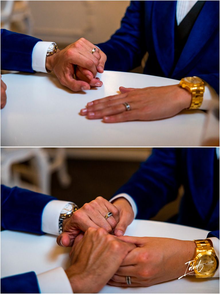 Close-up of hands with rings and gold watch signing papers, wearing navy blue suit sleeve and white dress shirt cuff.