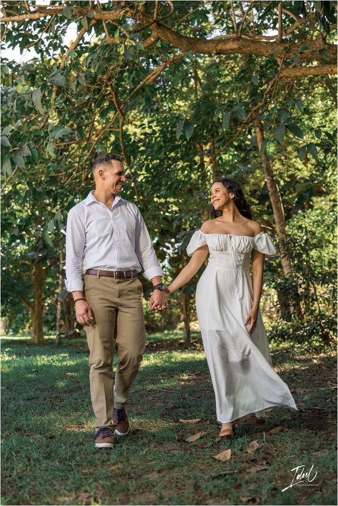A couple walks hand in hand through a sun-dappled garden, with her wearing a flowing white dress and him in khakis.