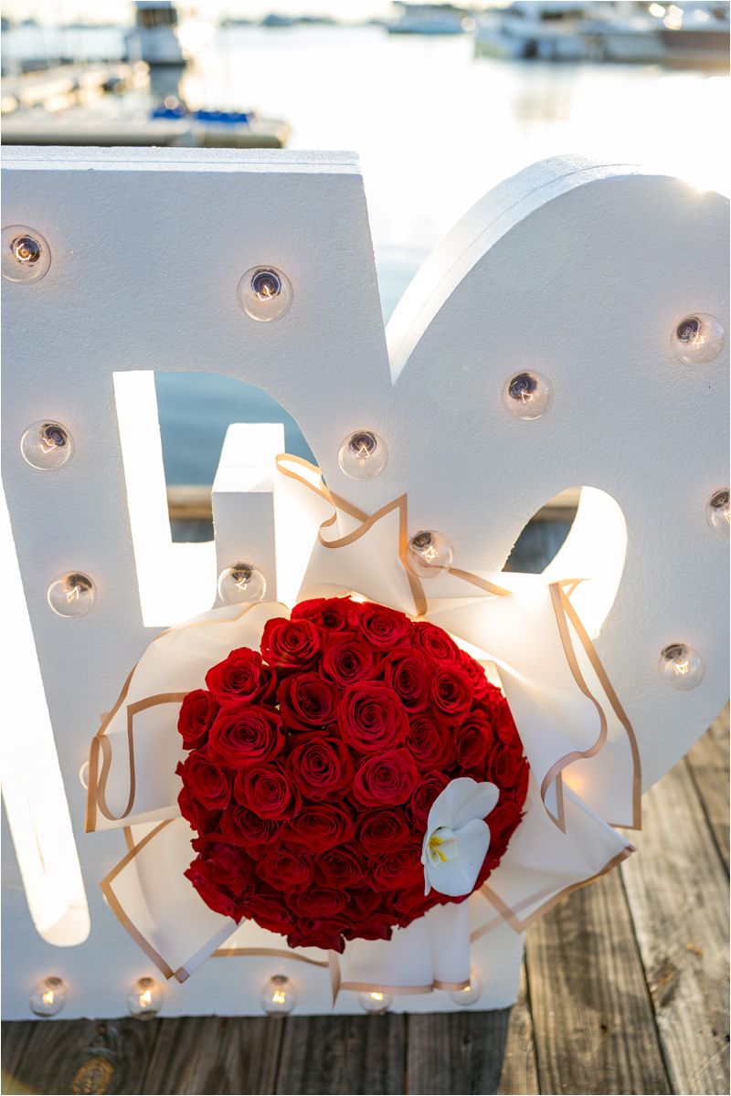Large illuminated white 'LOVE' letters with red rose bouquet and ribbon on a wooden dock by the water.