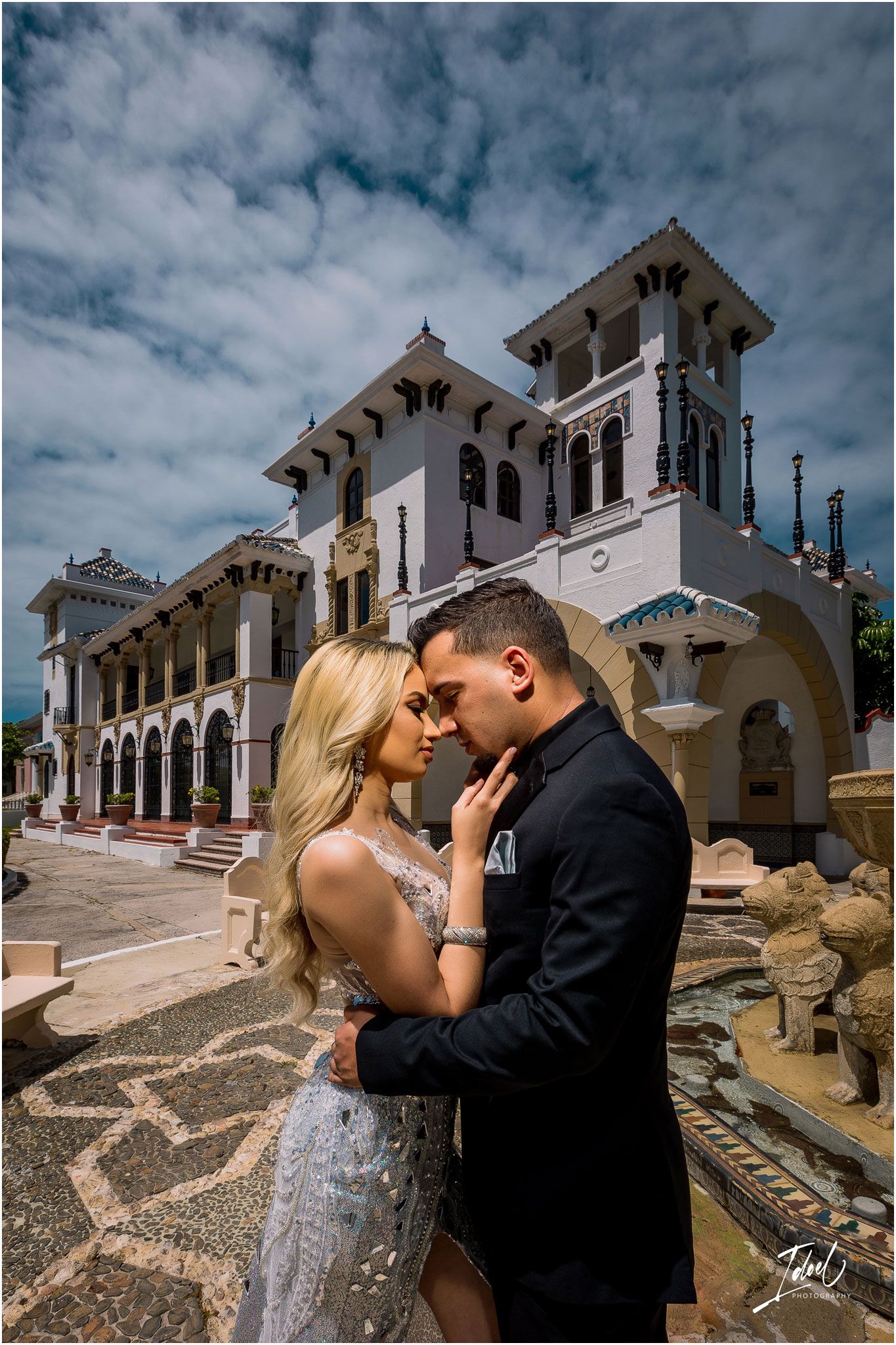 Intimate moment captured in front of Spanish colonial building with dramatic cloudy sky.