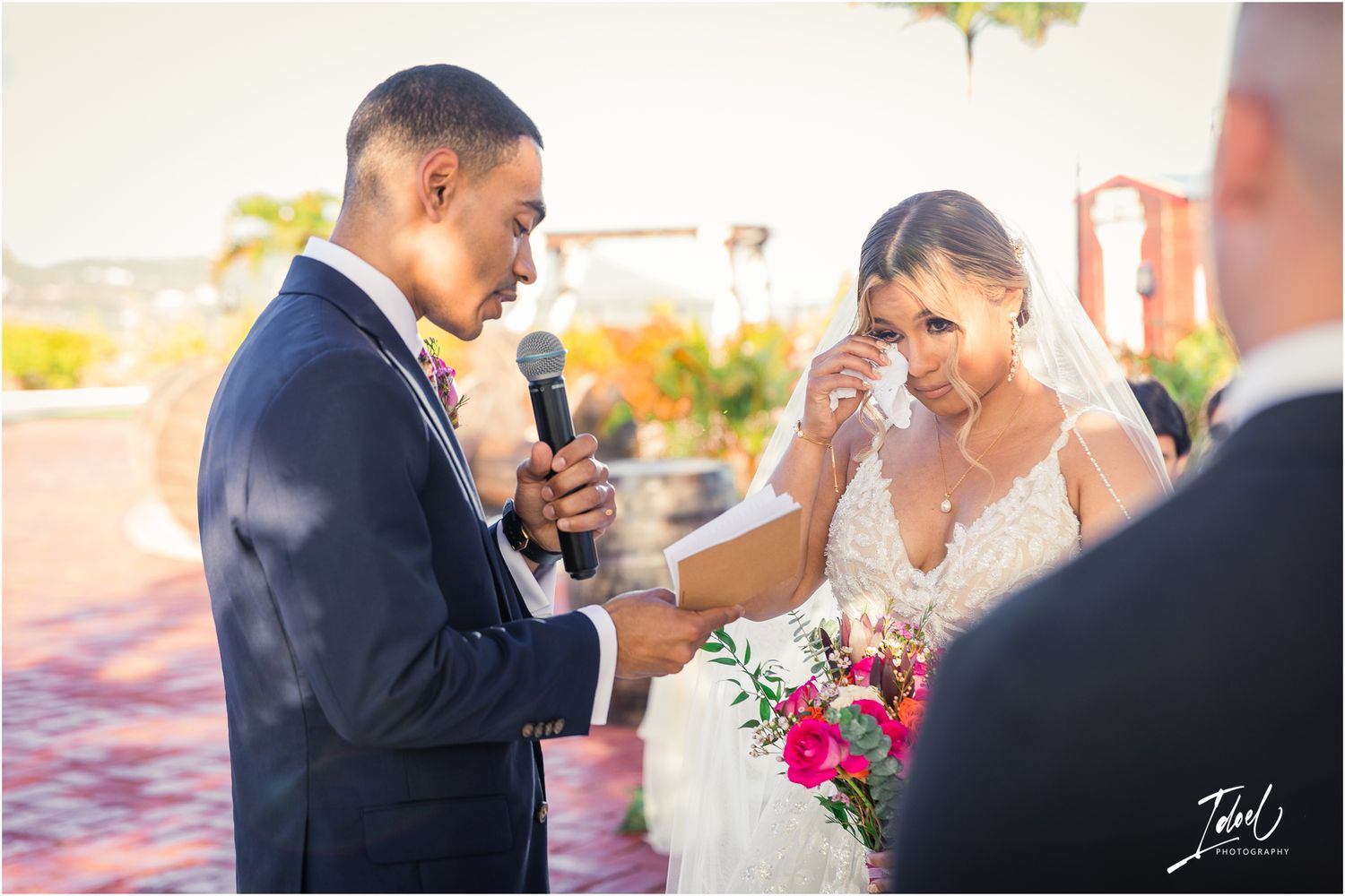 Emotional moment during wedding vows at an outdoor ceremony with pink floral bouquet.