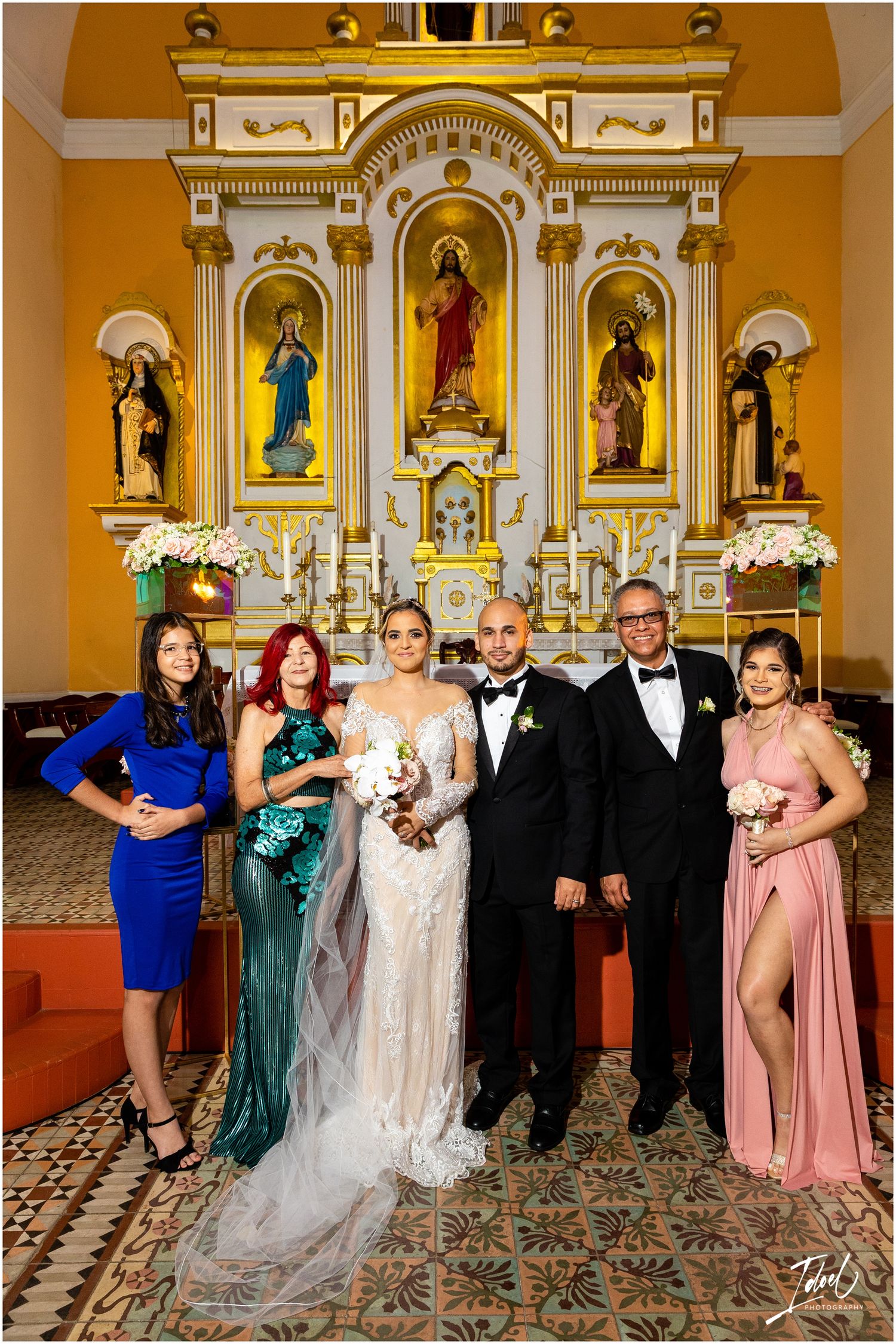 Small wedding party in formal attire poses in front of ornate church altar with religious iconography.